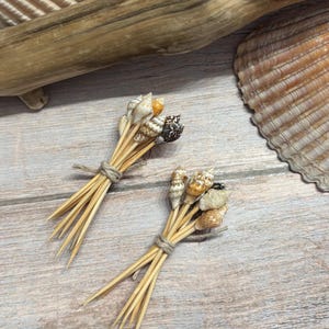 Puede incluir: Dos pequeños ramos de conchas atados con hilo. Cada ramo presenta una colección de pequeñas conchas coloridas unidas a brochetas de madera. El fondo incluye una concha grande y madera flotante sobre una superficie de madera clara.