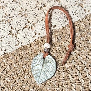 May include: A leaf-shaped pendant with a light green finish, detailed with veining, hangs from a brown leather cord. A white bead and silver-toned metal accents are at the top. The pendant is a nature-inspired accessory.