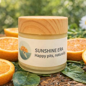 May include: A glass jar with a wooden lid, labelled "SUNSHINE ERA Happy pits, naturally." The jar is surrounded by orange halves and green leaves. A daisy is in the background. The jar contains a yellow cream.