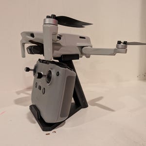 May include: A gray drone with black propellers and a matching remote control, mounted on a black stand. The drone's arms are extended, and the remote control has control sticks and buttons. The setup is designed for aerial photography and videography.