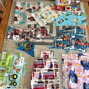 May include: A collection of assorted cotton fabric scraps featuring various character prints including Moana, Spider-Man, Batman, Paddington Bear, Finding Nemo, and John Deere tractors, arranged on a gray cutting mat with inch markings.