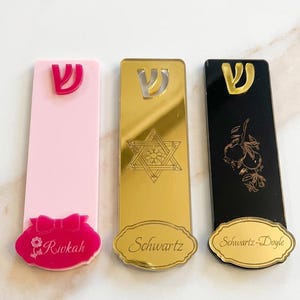 May include: Three decorative mezuzah cases in pink, gold, and black. The pink case has a bow and the name "Rivkah". The gold case has a Star of David and the name "Schwartz". The black case has a floral design and the name "Schwartz-Doyle".