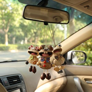 May include: Three novelty car air fresheners suspended from a car's rearview mirror. Each features an acorn-shaped head, open mouth, and decorative bow. The figures are brown, beige, and red, adding a touch of fun to the vehicle.