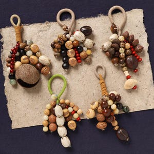 May include: Five handmade keychains featuring a variety of natural beads and seeds in shades of brown, beige, and red. Each keychain has a loop for hanging and a leather or cord accent. The keychains are arranged on a textured, off-white surface.