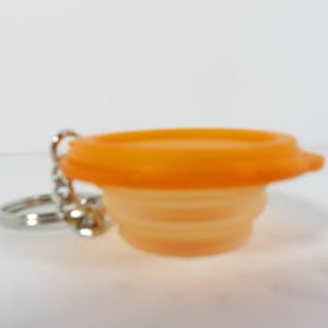 May include: An orange collapsible silicone cup with a matching lid, attached to a silver keychain. The cup is accordion-style, and the lid has a small tab for easy opening. The keychain is made of a chain and a split ring.