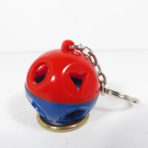 May include: A red and blue spherical keychain with geometric cutouts. The keychain has a silver chain and ring. The base is gold-colored. The sphere has a red top and a blue bottom.
