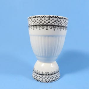 May include: White ceramic egg cup with a pedestal base and a wide, ribbed body. The rim and base are decorated with a black floral pattern. The egg cup is set against a light blue background.