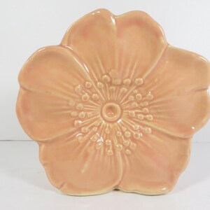 May include: A peach-colored ceramic dish shaped like a five-petaled flower. The surface features a glossy finish with raised, radiating lines and small, rounded bumps in the center, mimicking the appearance of stamens on a natural blossom.