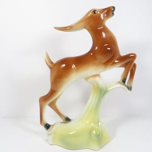May include: A glossy ceramic figurine of a leaping deer with a brown body, white underbelly, and dark brown hooves. The deer is supported by a pale yellow and green base that mimics a tree stump, featuring a smooth, reflective finish throughout.