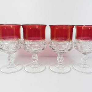 May include: Four vintage wine glasses with ruby red rims and clear, patterned bowls. The stems are clear and have a decorative design. The glasses are arranged in a row against a white background. The glasses are likely made of glass.