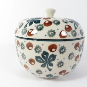 May include: A round ceramic lidded jar shaped like an apple with a cream-colored base. The surface features a hand-painted pattern of dark green chestnut leaves, brown chestnuts, and small blue dots scattered across the entire exterior.