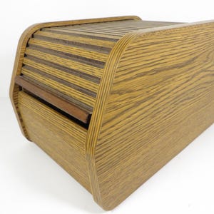 May include: A rectangular bread box featuring a light brown wood grain finish and a curved top. The unit includes a sliding tambour door with horizontal slats that retract to reveal the interior compartment, constructed with visible layered plywood edges.