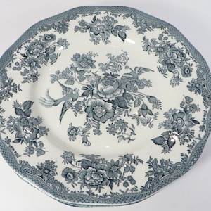 May include: A round ceramic plate featuring a blue and white transferware pattern. The design includes central floral arrangements with blooming flowers and two birds, surrounded by a decorative border of intricate lattice and floral motifs.