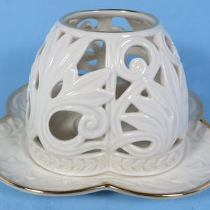 May include: A white ceramic candle holder with a floral design. The holder has a decorative, openwork design with leaf and swirl patterns. It sits on a matching saucer with a gold rim. The background is a solid blue.