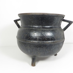 May include: A black cast iron cauldron with a rounded body, a wide open top, and two triangular side handles. The vessel features a textured surface and stands on three short, sturdy legs, showcasing a dark, weathered metallic finish.