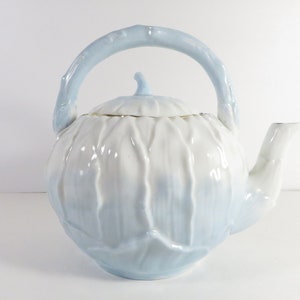 May include: A white and light blue ceramic teapot shaped like a cabbage with a twig-like handle.