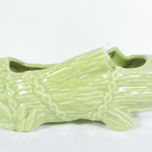 May include: A light green ceramic planter shaped like a log, with a textured surface and a rope design. The planter has two openings for plants and is marked "USA" on the side. The overall design is rustic and decorative.