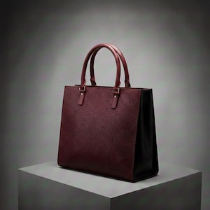 Puede incluir: Bolso tote burdeos con un patrón geométrico en el panel frontal. El bolso tiene dos asas superiores y forma rectangular. Los laterales del bolso son negros y las asas son de color burdeos a juego.