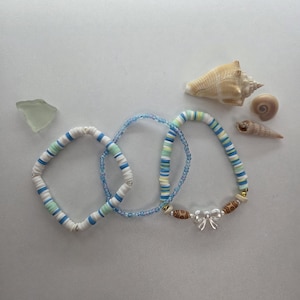 Op de afbeelding: Drie armbanden met blauwe, witte en groene kralen, samen met schelpen en zeeglas. Eén armband heeft een strikbedel. De armbanden zijn gemaakt van kleine, platte kralen. Het thema is kust en strand.