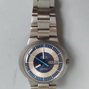 May include: Silver-toned Omega Genève Dynamic automatic wristwatch. The watch features a blue and silver dial with a day-date display and a silver-toned metal band. The watch face has the text "Genève Dynamic" and "SAB 25".