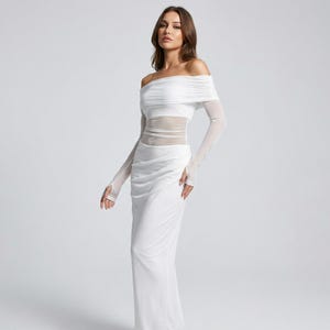 May include: A white off-the-shoulder dress with long, sheer sleeves and a fitted bodice. The dress features a draped neckline and a floor-length skirt. The sleeves extend to the knuckles, with a finger loop. The dress is made of a soft, flowing fabric.