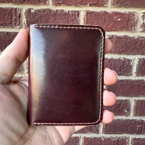 May include: A rectangular dark burgundy leather wallet featuring a smooth, polished finish and visible light brown stitching along the edges. The wallet has rounded corners and a vertical orientation, showcasing a clean, minimalist design.