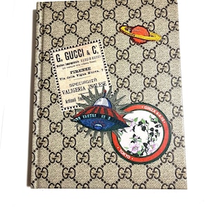 May include: A notebook with a repeating brown pattern of interlocking Gs. The cover features patches with text, a planet, and a UFO. The patches include the text "G. GUCCI & C." and "TOKYO".