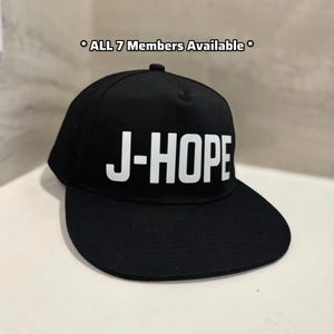 Puede incluir: Gorra de béisbol negra con el nombre "J-HOPE" en letras blancas y en negrita en la parte delantera. El texto "* ALL 7 Members Available *" está encima de la gorra. La gorra tiene una visera plana y una corona estructurada.
