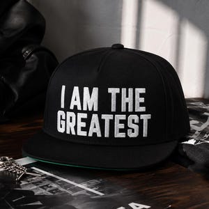 May include: A black baseball cap with a flat brim and the white embroidered text I AM THE GREATEST centered on the front panel. The cap features a structured crown with a top button and a green underside visible on the edge of the flat brim.