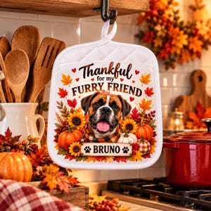 May include: A white square potholder with a hanging loop, featuring a graphic of a Boxer dog wearing a plaid scarf, surrounded by autumn leaves, pumpkins, and sunflowers. The text reads Thankful for my FURRY FRIEND and BRUNO.