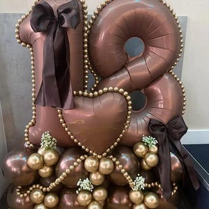Pode incluir: Um arranjo festivo de balões castanhos formando o número 18, acentuado com bordas de contas douradas e laços castanhos escuros. Um balão em forma de coração está no centro, e balões dourados e castanhos menores completam a exibição. Pequenas flores brancas.