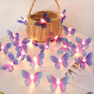 Puede incluir: Una cadena de luces decorativa con adornos en forma de mariposa. Las mariposas tienen un degradado de tonos rosa y azul, con pequeñas luces en el centro. Una cesta de mimbre se encuentra en el fondo.