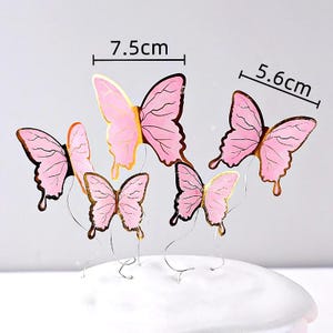 May include: Pink butterfly decorations with gold trim, measuring 7.5cm and 5.6cm. These decorative butterflies are attached to thin wire stems, perfect for cake toppers or craft projects. The butterflies have a delicate, layered design.