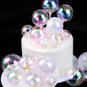 Puede incluir: Un pastel blanco decorado con adornos iridiscentes en forma de burbujas. Los adornos varían en tamaño y están sujetos al pastel con tallos rosas. Algunas burbujas tienen un brillo nacarado, reflejando múltiples colores.