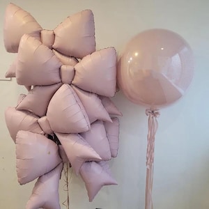 Könnte beinhalten: Eine Ansammlung von rosa Schleifenballons und einem großen runden Ballon. Die Schleifenballons sind gestapelt, während der runde Ballon mit einem Band gebunden ist. Die Ballons haben eine weiche, pastellrosa Farbe, ideal für eine Party oder Feier.