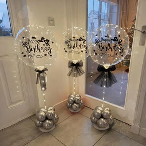 Puede incluir: Tres globos transparentes con el texto "Happy Birthday" y elementos decorativos. Cada globo está iluminado con pequeñas luces y adornado con un lazo negro. Los globos están sostenidos por grupos de globos plateados.