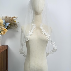 Könnte beinhalten: Ein durchsichtiger weißer Brautschleier mit zarten floralen Spitzenbesatz. Der Schleier wird auf einer Schaufensterpuppe präsentiert, die sein fließendes Design zeigt. Die Spitzendetails verleihen diesem Hochzeitsaccessoire einen Hauch von Eleganz.