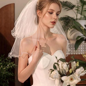 May include: A white two-tier bridal veil featuring a delicate rolled edge finish. The sheer tulle fabric is layered to create volume, attached to a hair comb, and worn with a strapless white gown while holding a bouquet of white magnolia flowers.