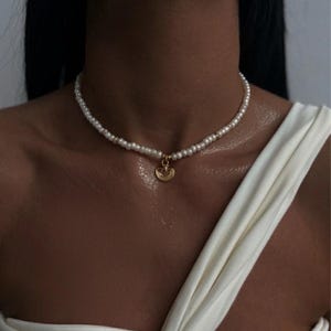 Könnte beinhalten: Eine zarte Perlenkette mit einem goldenen Anhänger. Die Halskette besteht aus kleinen, runden Perlen, die zu einem Choker verarbeitet sind. Der goldene Anhänger ist ein kleines, kreisförmiges Design mit detaillierten Mustern. Die Halskette wird über einem weißen Oberteil getragen.