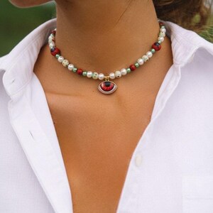 K&ouml;nnte beinhalten: Eine Perlenhalskette als Choker mit wei&szlig;en, hellgr&uuml;nen und roten runden Perlen. Die Kette hat einen zentralen Anh&auml;nger in Form eines Auges mit roter Iris und schwarzer Pupille, der an einer kleinen goldfarbenen Metall&ouml;se h&auml;ngt.
