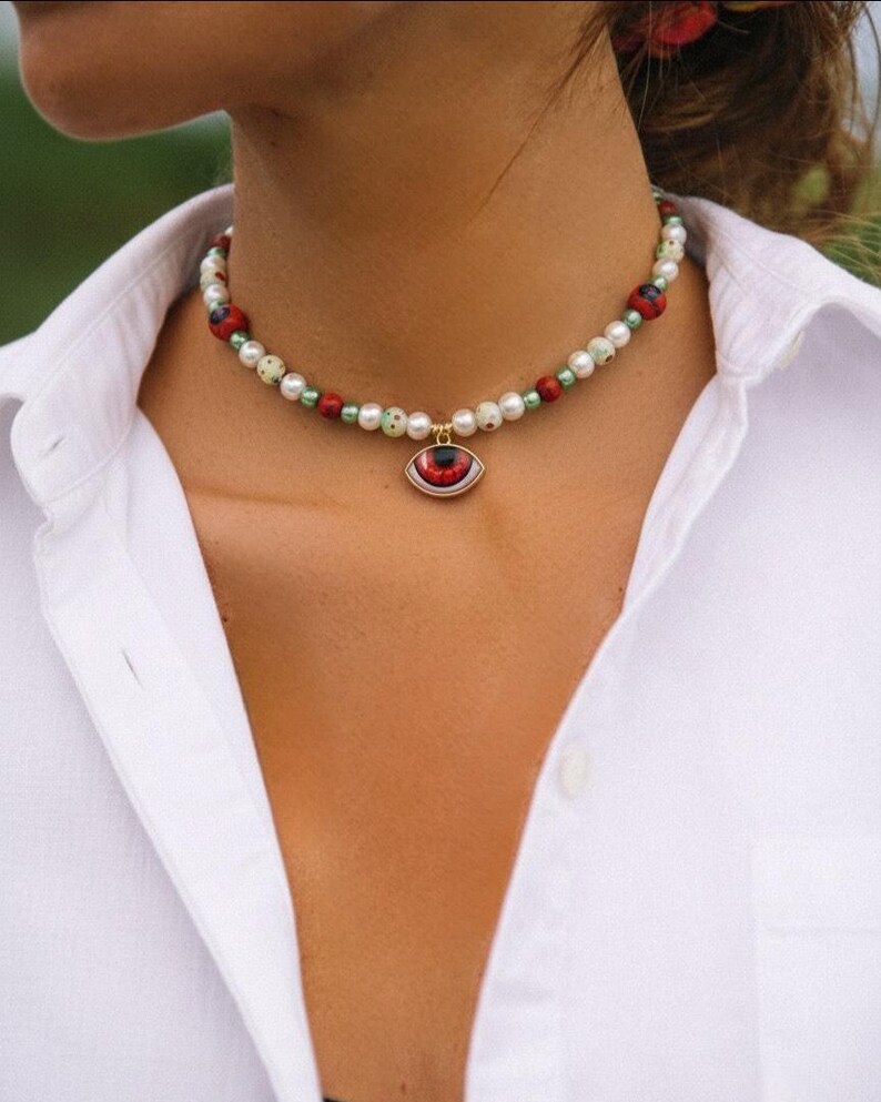 K&ouml;nnte beinhalten: Eine Perlenhalskette als Choker mit wei&szlig;en, hellgr&uuml;nen und roten runden Perlen. Die Kette hat einen zentralen Anh&auml;nger in Form eines Auges mit roter Iris und schwarzer Pupille, der an einer kleinen goldfarbenen Metall&ouml;se h&auml;ngt.