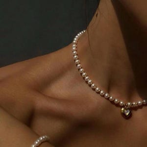 Könnte beinhalten: Eine Perlenkette mit einem goldenen Herzanhänger. Die Kette besteht aus kleinen, runden weißen Perlen und goldenen Perlen. Der Herzanhänger hat einen klaren, irisierenden Stein in der Mitte. Ein passendes Perlenarmband ist sichtbar.