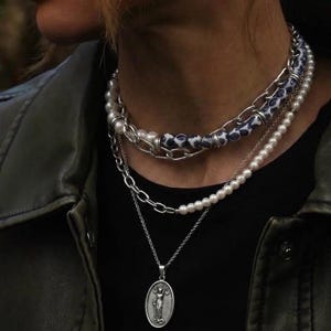 May include: A layered necklace set featuring a top strand of white pearls and blue-and-white patterned beads mixed with silver-tone chain links, a middle strand of white pearls and silver-tone chain, and a bottom silver-tone chain with an oval pendant.