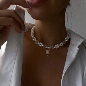 Könnte beinhalten: Eine zarte Halskette mit einer doppelten Reihe aus weißen Perlen und klaren, rosafarbenen Perlen. Ein goldfarbener Anhänger mit Sonnenmuster hängt in der Mitte. Die Halskette wird über einem weißen Hemd getragen.