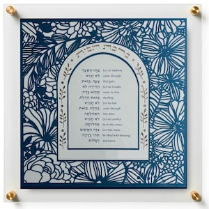 May include: A square wall art piece featuring a blue floral laser-cut overlay on a white background. The center displays an arched frame with Hebrew and English text: Let no sadness come through this gate. Let no trouble come to this dwelling. Let no fear come through this door. Let no conflict be in this place. Let this home be filled with blessing and peace. The piece is mounted between two clear acrylic panels with gold-toned hardware at the corners.