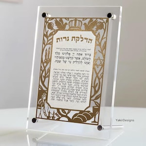 May include: Clear acrylic plaque with gold text. The text is in Hebrew and includes the words "Hadlakat Nerot." The plaque has a decorative border with floral and crown motifs. The plaque is supported by a clear stand.