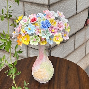 以下が含まれることがあります： 手作りの花束が入ったカラフルなセラミック花瓶。花はピンク、青、黄色、緑の色合いです。花瓶はテクスチャ加工されたパステルカラー仕上げで、濃い木製のテーブルの上に置かれています。