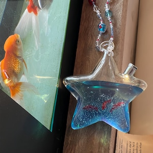May include: A star-shaped glass vessel filled with blue liquid and small red fish-shaped inserts, featuring a metallic spout and a decorative hanging loop adorned with multicolored glass beads, positioned next to a fish tank with live goldfish.