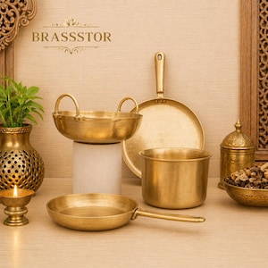 May include: A collection of brass cookware and serving dishes. The set includes a wok-style pan, a frying pan, a saucepan, a serving bowl, and a lidded container. The pieces are arranged on a light-colored surface with the brand name "BRASSSTOR" visible.