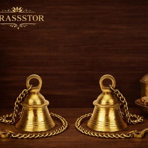 Peut inclure: Deux cloches en laiton doré avec chaînes et crochets sur une surface en bois. Les cloches sont au premier plan, avec un vase décoratif, un petit bougeoir et un bol d'épices en arrière-plan. La marque "BRASSSSTOR" est en haut.
